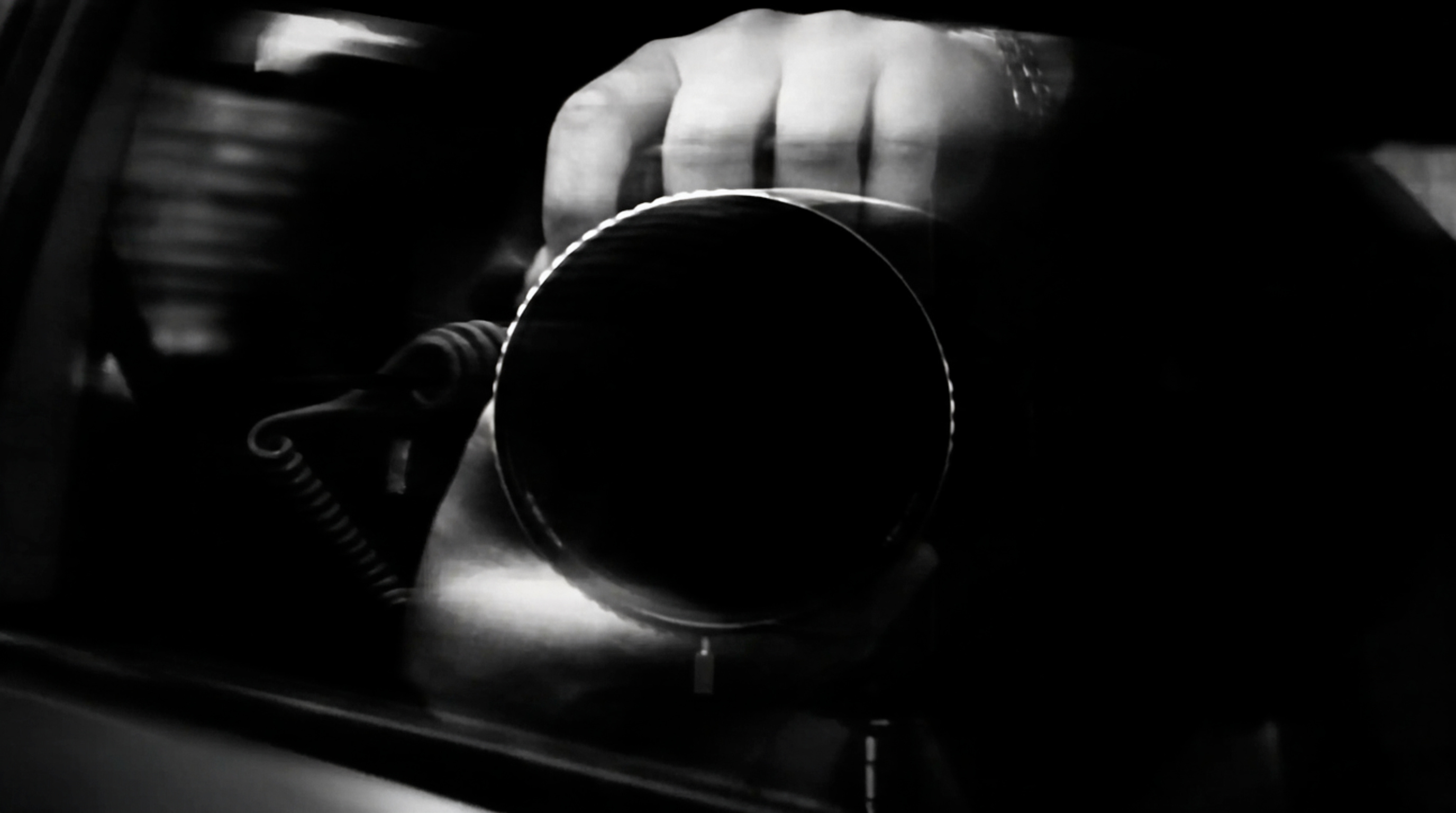 Black and white close-up of a hand holding a camera, lens pointed at the viewer